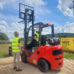 Instruktor w żółtej kamizelce obserwuje kursanta obsługującego czerwony wózek widłowy spalinowy na placu manewrowym, widoczny baner reklamowy z informacjami o kursach w tle.