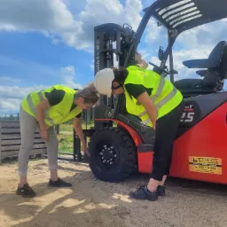 Dwie osoby w kamizelkach odblaskowych sprawdzają opony czerwonego wózka widłowego na piaszczystym terenie, widoczna naklejka z danymi kontaktowymi firmy.