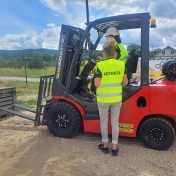 TECHNICAL ANETA KASPRZAK - Instruktor z kursantem na wózku widłowym na placu manewrowym, w tle zalesione wzgórza i błękitne niebo z chmurami.
