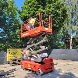 TECHNICAL ANETA KASPRZAK - Czerwony podnośnik nożycowy Maniaccess 100 XEL z pracownikiem w kasku i kamizelce na platformie, na tle zieleni drzew i baneru reklamowego kursów na wózki widłowe.