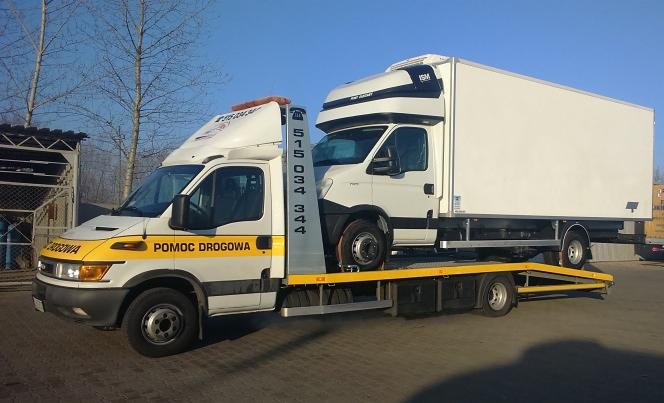 Laweta z napisem 'Pomoc Drogowa' przewozi biały samochód dostawczy na platformie. Widoczne numery kontaktowe na lawetcie. Błękitne niebo w tle.