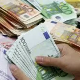 Dłoń trzymająca plik banknotów euro na tle rozłożonych pieniędzy różnych nominałów, w tym 500, 100 i 50 euro.