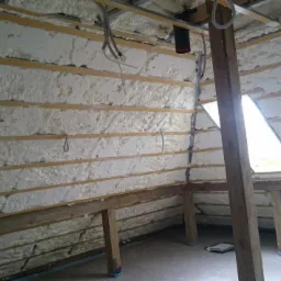 Poddasze z widoczną strukturą drewnianą i ociepleniem pianką PUR, z przewodami elektrycznymi w górnej części ściany i oknem dachowym.