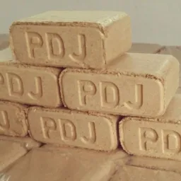 Ułożone w piramidę brykiety drzewne z wytłoczonym napisem 'PDJ'. Brykiety leżą na palecie zabezpieczonej folią. Jasne, neutralne tło.