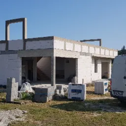 Niedokończony budynek z białych bloczków silikatowych z widocznymi schodami wewnętrznymi, palety z materiałami budowlanymi i biały samochód dostawczy w tle.