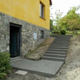 Betonowe schody prowadzące od wejścia do budynku z żółtą elewacją, widoczne ślady świeżego betonu i ziemi po budowie.