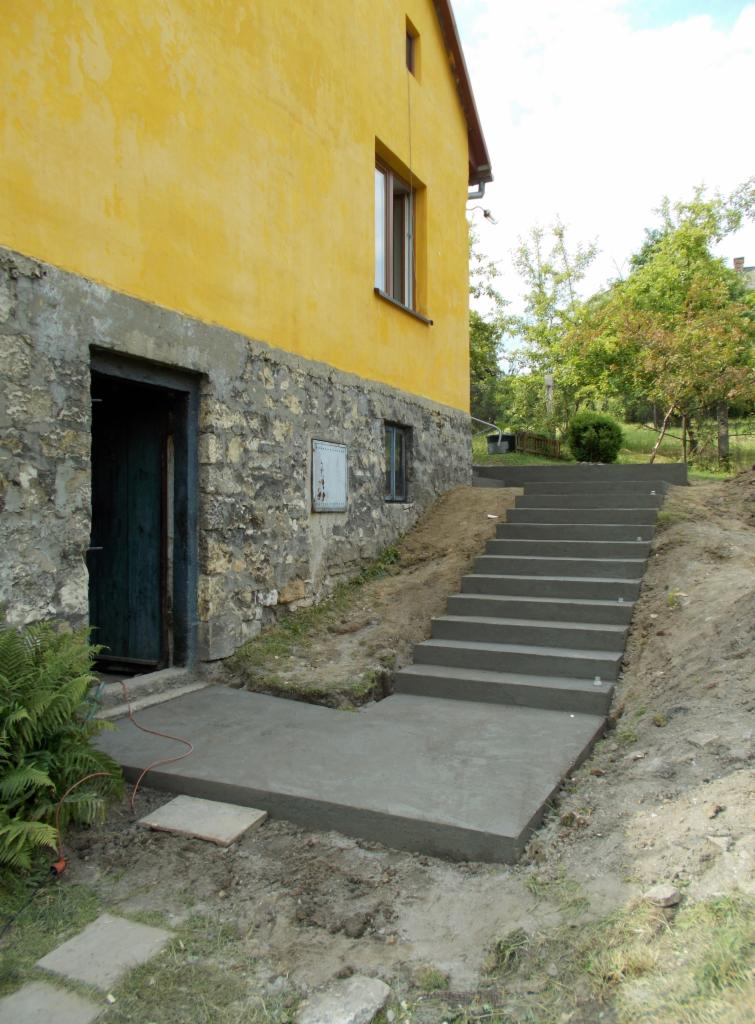 Betonowe schody prowadzące od wejścia do budynku z żółtą elewacją, widoczne ślady świeżego betonu i ziemi po budowie.