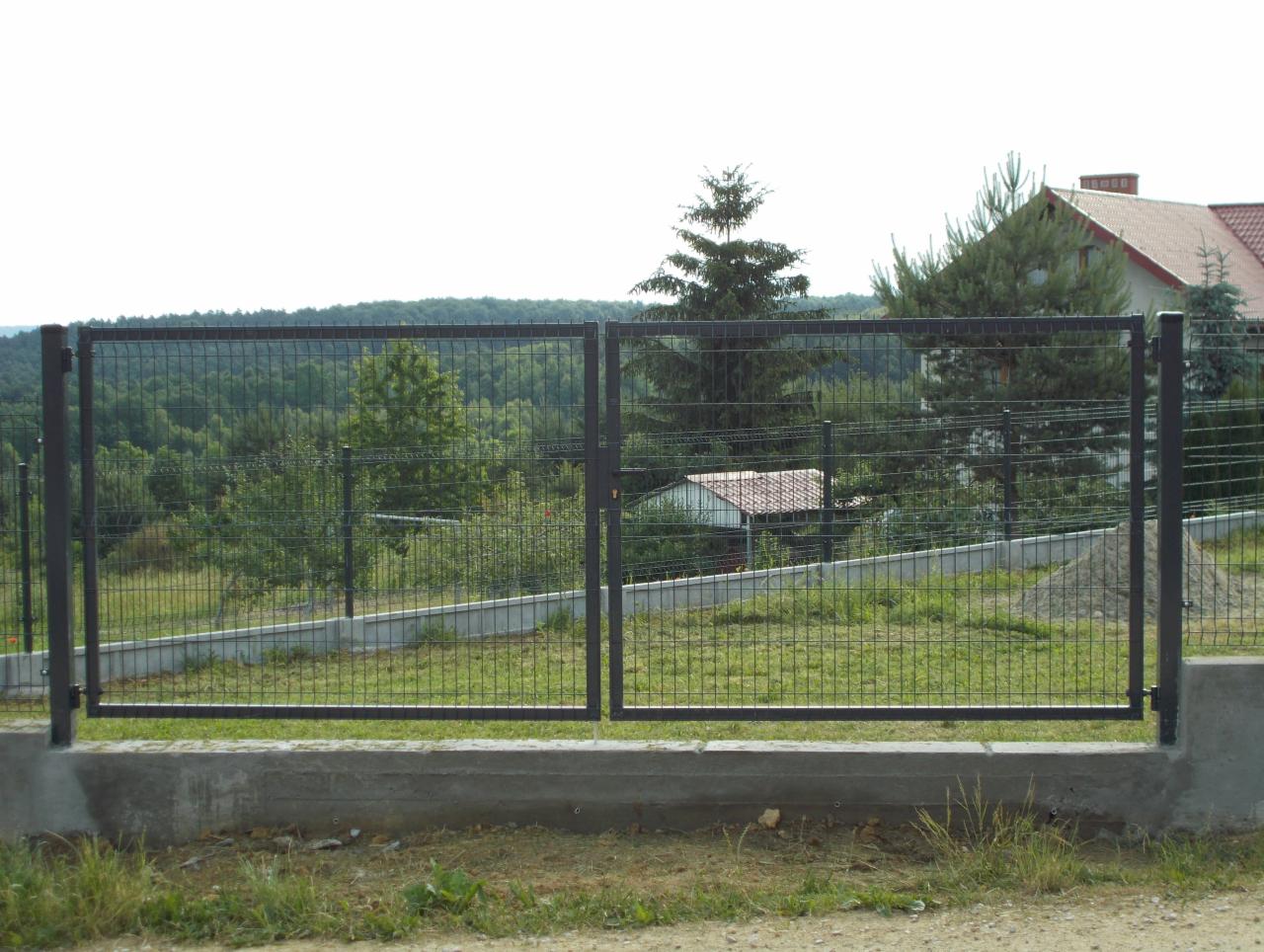 Metalowa brama dwuskrzydłowa z siatki na betonowej podmurówce, widok na zalesione wzgórze i dom z czerwonym dachem w tle.
