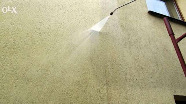 Czyszczenie elewacji budynku za pomocą myjki ciśnieniowej. Strumień wody usuwa zabrudzenia z teksturowanej ściany zewnętrznej w słoneczny dzień.