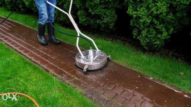 Czyszczenie kostki brukowej myjką ciśnieniową z rotacyjną dyszą; widoczne mokre, czyste fragmenty kostki i osoba w gumowych butach obsługująca urządzenie.