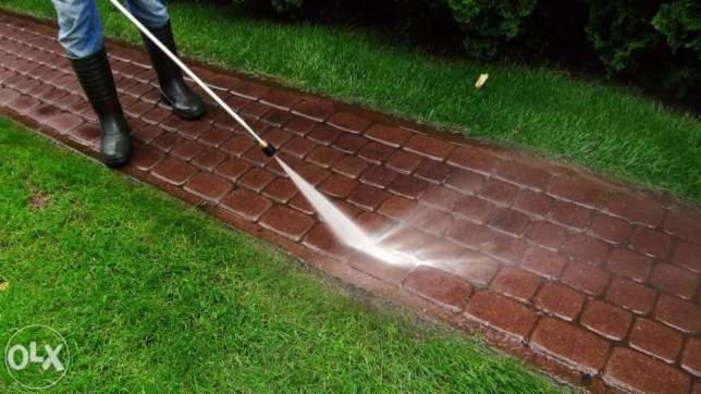 Czyszczenie kostki brukowej myjką ciśnieniową, woda pod wysokim ciśnieniem usuwa zabrudzenia z czerwonej kostki w otoczeniu zielonej trawy.