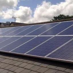 Rząd paneli słonecznych, w srebrnej ramie, zamontowany na ciemnoszarym, spadzistym dachu pokrytym dachówką bitumiczną, z anteną telewizyjną i budynkami w tle, pod błękitnym niebem z białymi chmurami.