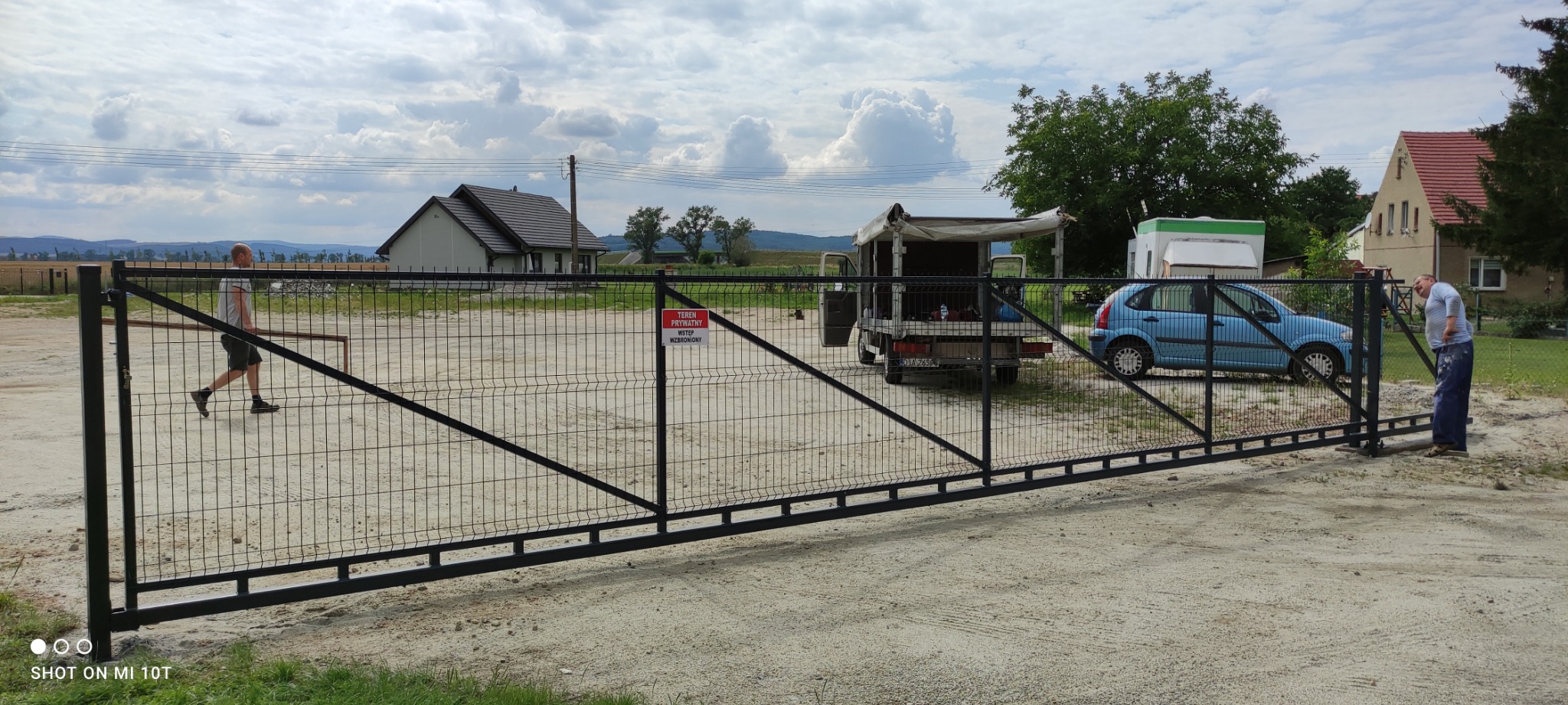 Czarna brama przesuwna z paneli ogrodzeniowych, widoczna z perspektywy placu budowy, z pracownikiem malującym jej fragment, w tle samochód dostawczy i domy.