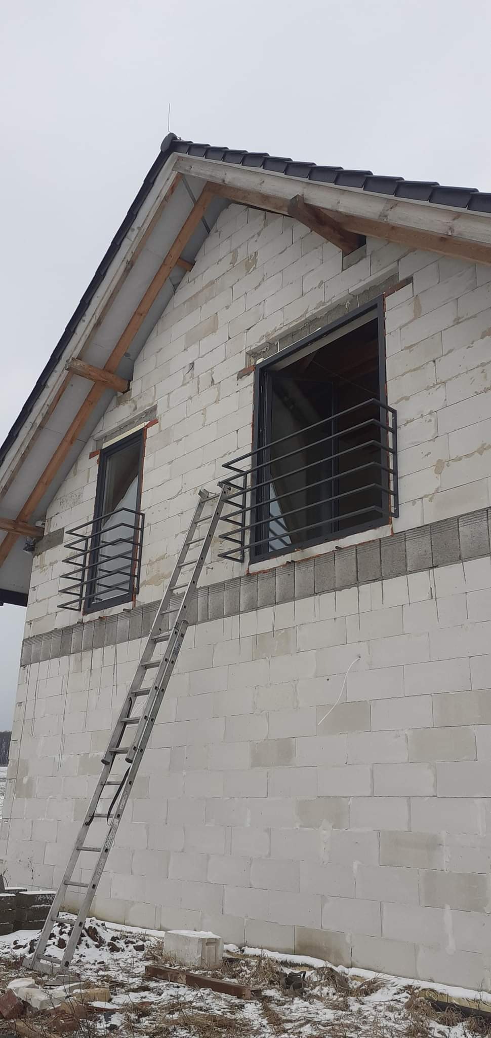 Surowe okna z prostymi metalowymi balustradami na ścianie domu z białych bloczków, drabina oparta o ścianę, widok z dołu, zimowy krajobraz w tle.