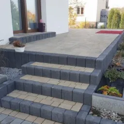 Schody i taras wyłożone kostką brukową w odcieniach szarości i beżu, z elementami małej architektury ogrodowej i roślinnością ozdobną.