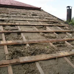Ocieplenie poddasza wełną mineralną między krokwiami. Widoczne fragmenty wełny i drewniana konstrukcja dachu. Prace ociepleniowe w toku, widok z góry.