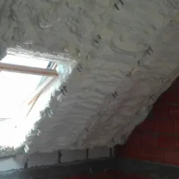 Ocieplenie poddasza pianką z widocznym oknem dachowym i fragmentem ściany z czerwonej cegły. Widoczne elementy montażowe pianki.