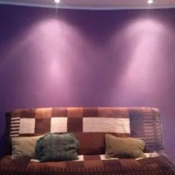 Fioletowa ściana oświetlona dwoma punktowymi lampami sufitowymi, nad brązowo-białą sofą z poduszkami.