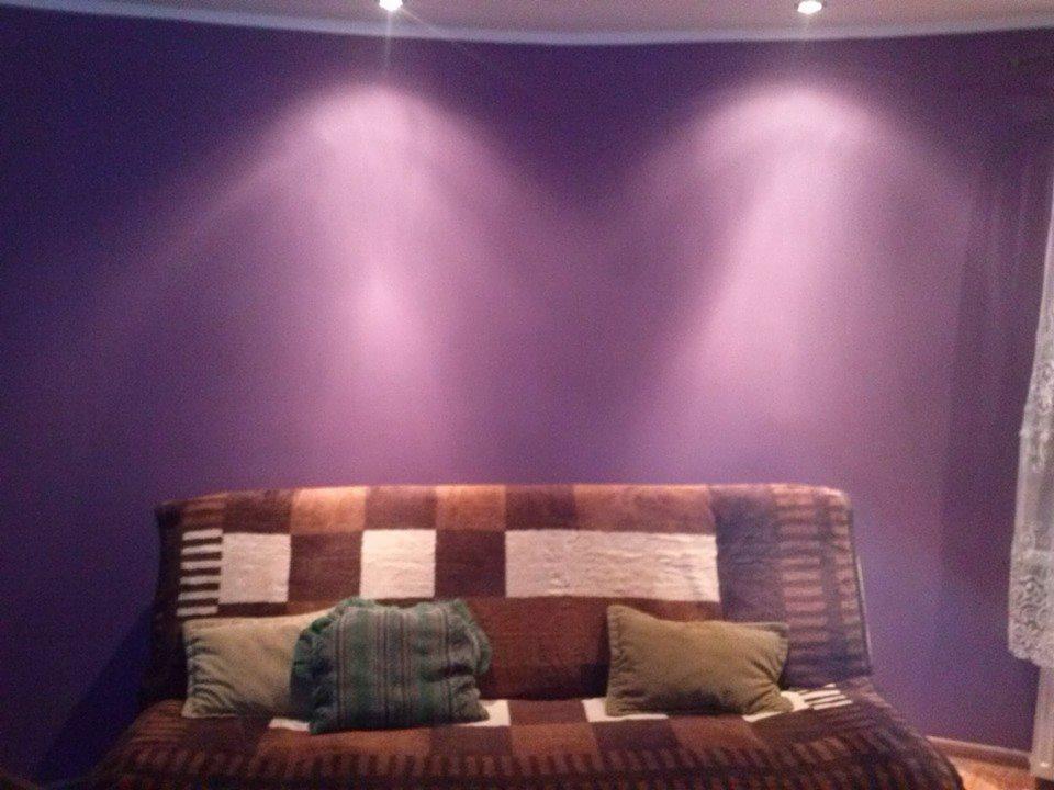 Fioletowa ściana oświetlona dwoma punktowymi lampami sufitowymi, nad brązowo-białą sofą z poduszkami.