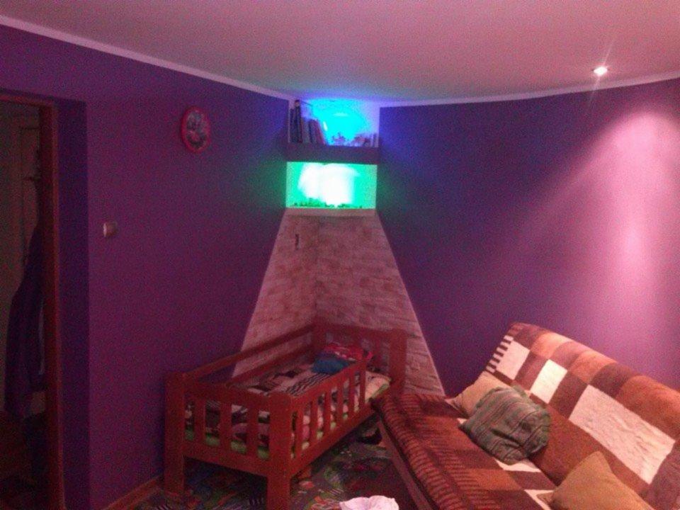 Pokój z fioletowymi ścianami, akwarium podświetlonym na zielono i niebiesko, obok łóżeczko dziecięce i kanapa z pledem w kratę.