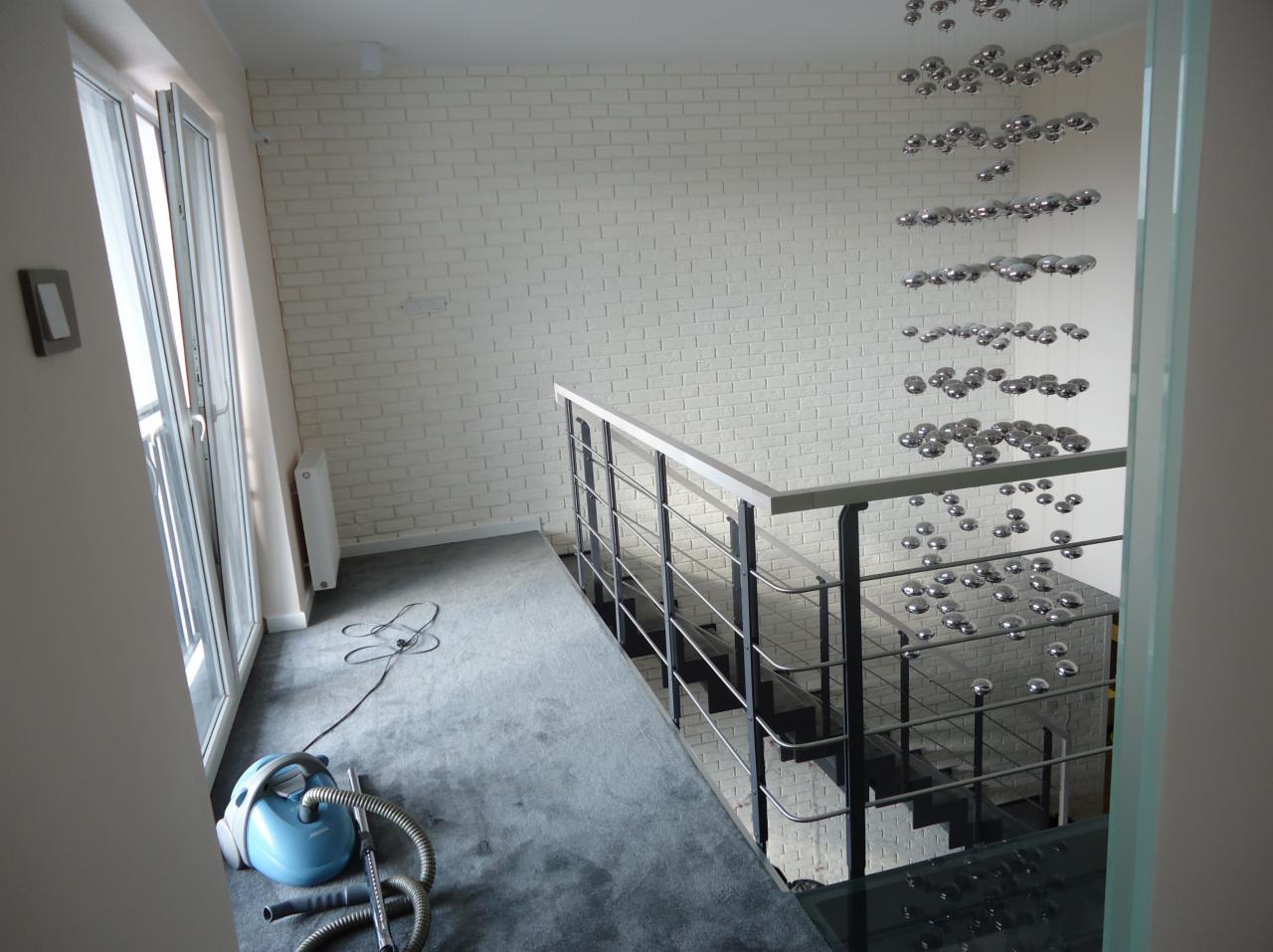 Wnętrze po remoncie: biała ceglana ściana, szare schody z metalową balustradą, niebieski odkurzacz. Widoczny element dekoracyjny - srebrne kule.