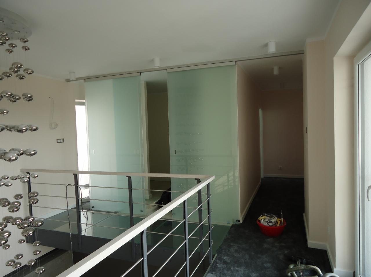 Nowoczesne, przesuwne szklane drzwi z matowym wykończeniem oddzielają przestrzeń. Widoczne balustrady, żyrandol i akcesoria malarskie w czerwonym wiadrze.