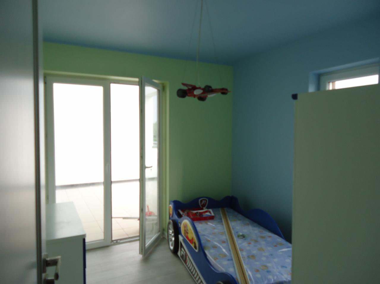 Pokój dziecięcy z łóżkiem w kształcie samochodu, świeżo pomalowane ściany w dwóch odcieniach zieleni i błękitu, z otwartym oknem balkonowym i lampą w kształcie samolotu.