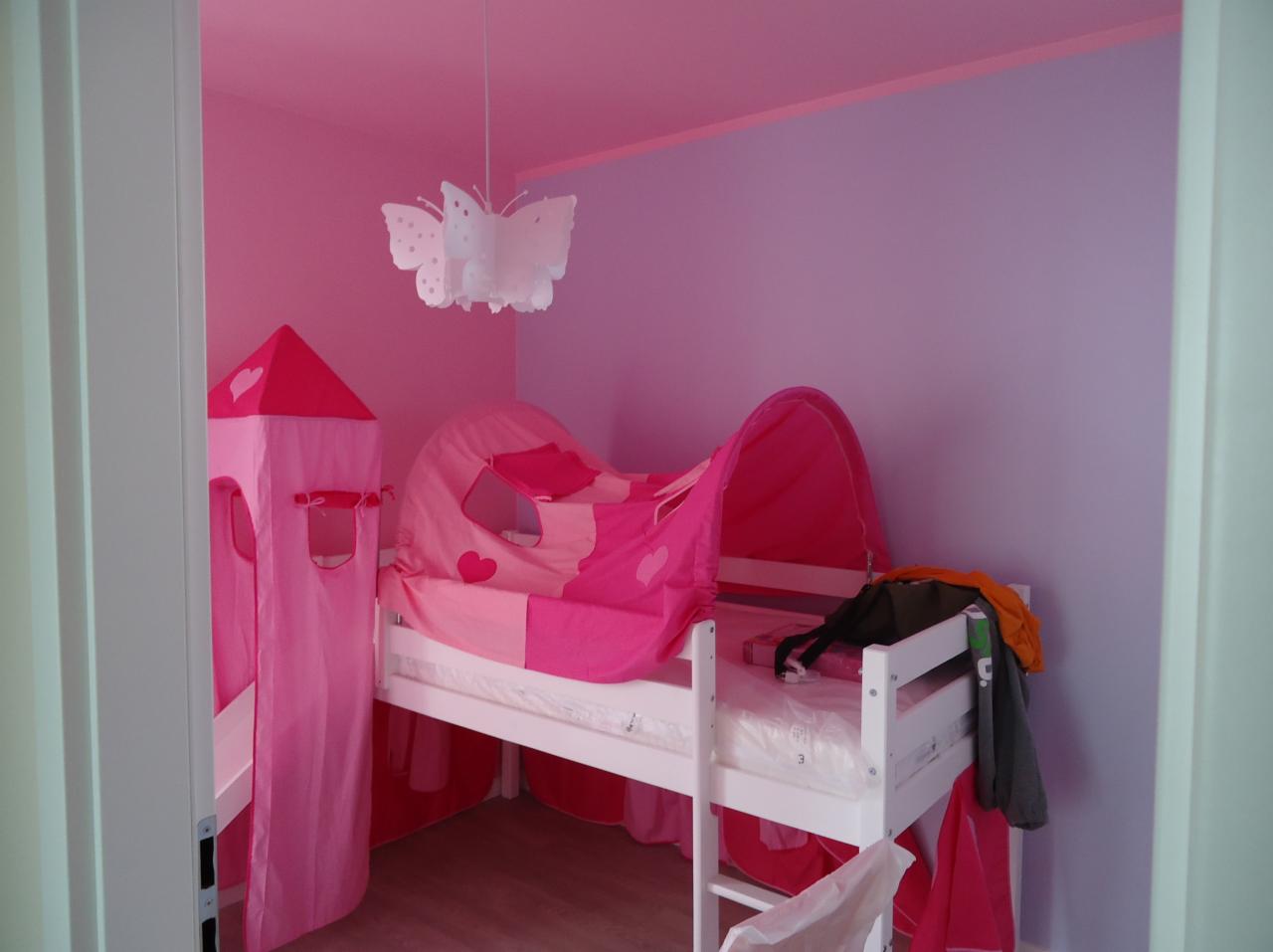 Pokój dziecięcy w odcieniach różu i fioletu z białym łóżkiem z baldachimem i wieżą. Na ścianie wisi lampa w kształcie motyla. Całość tworzy bajkową atmosferę.