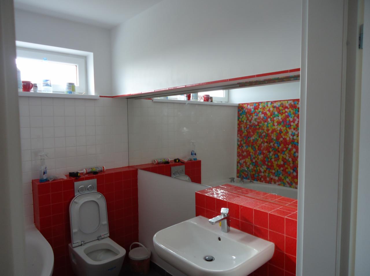 Wykończona łazienka z czerwonymi akcentami: czerwone płytki na ścianach, umywalka z baterią, toaleta i duże lustro odbijające kolorową ścianę nad wanną.