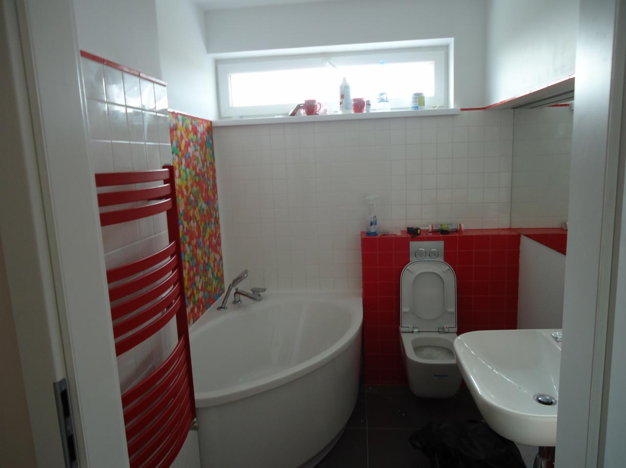 Biała wanna, umywalka i toaleta w wyremontowanej łazience z czerwonym grzejnikiem drabinkowym, mozaiką i akcesoriami. Okno nad wanną. Widok zza drzwi.
