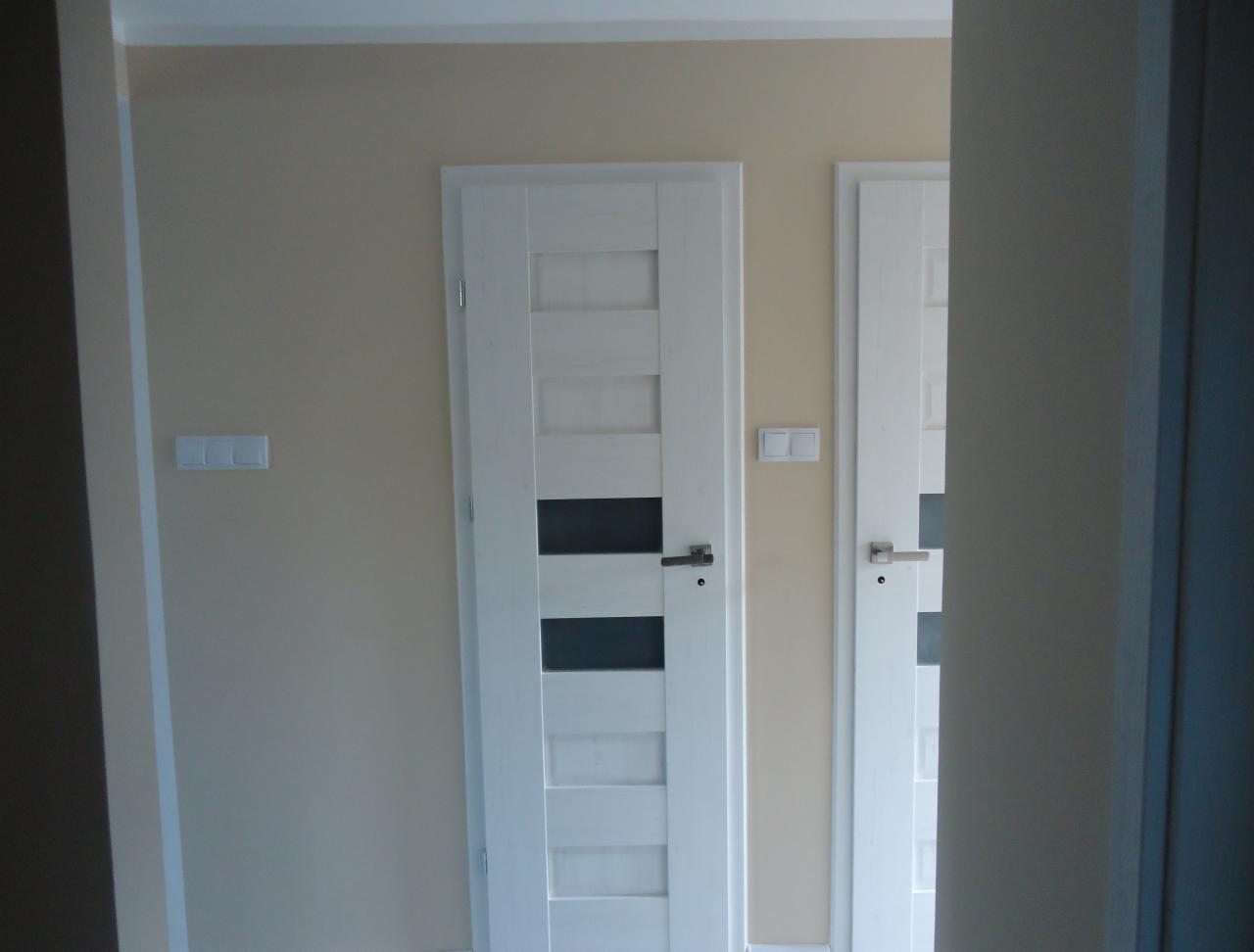 Dwie białe, nowoczesne drzwi z przeszkleniami, zamontowane w beżowej ścianie z widocznym włącznikiem. Kadrowanie zza framugi, widok na korytarz.