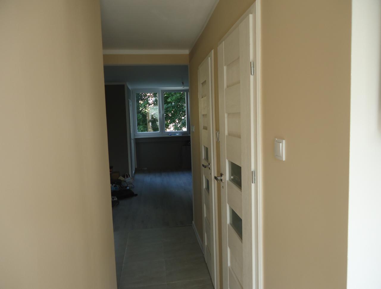 Jasny korytarz po remoncie z dwiema białymi, przeszklonymi drzwiami po prawej stronie i widokiem na pokój z oknem na końcu. Ściany pomalowane na beżowo.