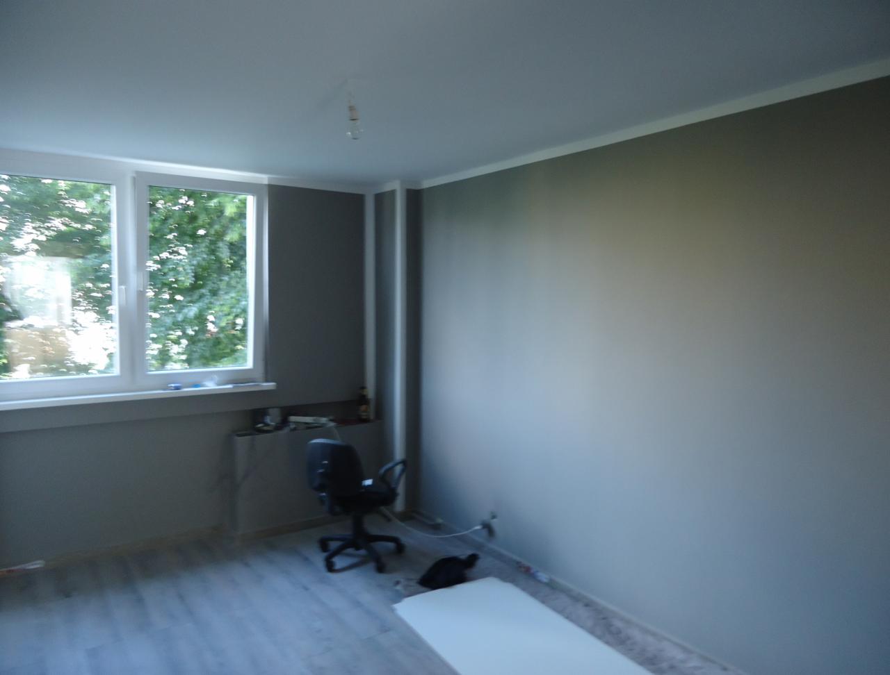 Pomieszczenie po malowaniu ścian na szaro, z widocznym oknem, krzesłem biurowym i fragmentem podłogi. Świeżo pomalowane ściany o jednolitej barwie.