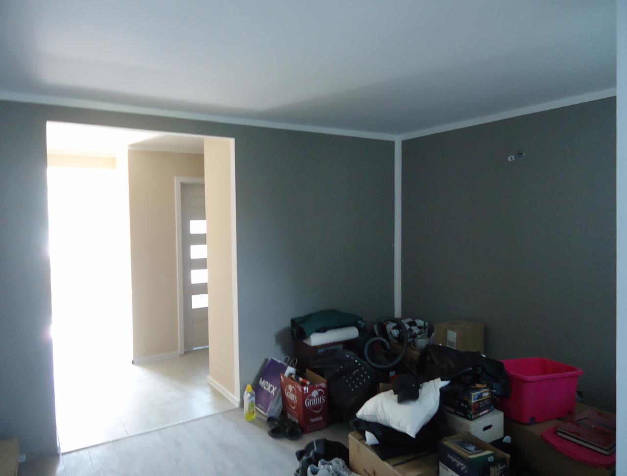 Pokój przed remontem: szare ściany, białe listwy, nagromadzone przedmioty. Widoczne drzwi wejściowe i fragment jasnego pomieszczenia. Chaos przed metamorfozą.