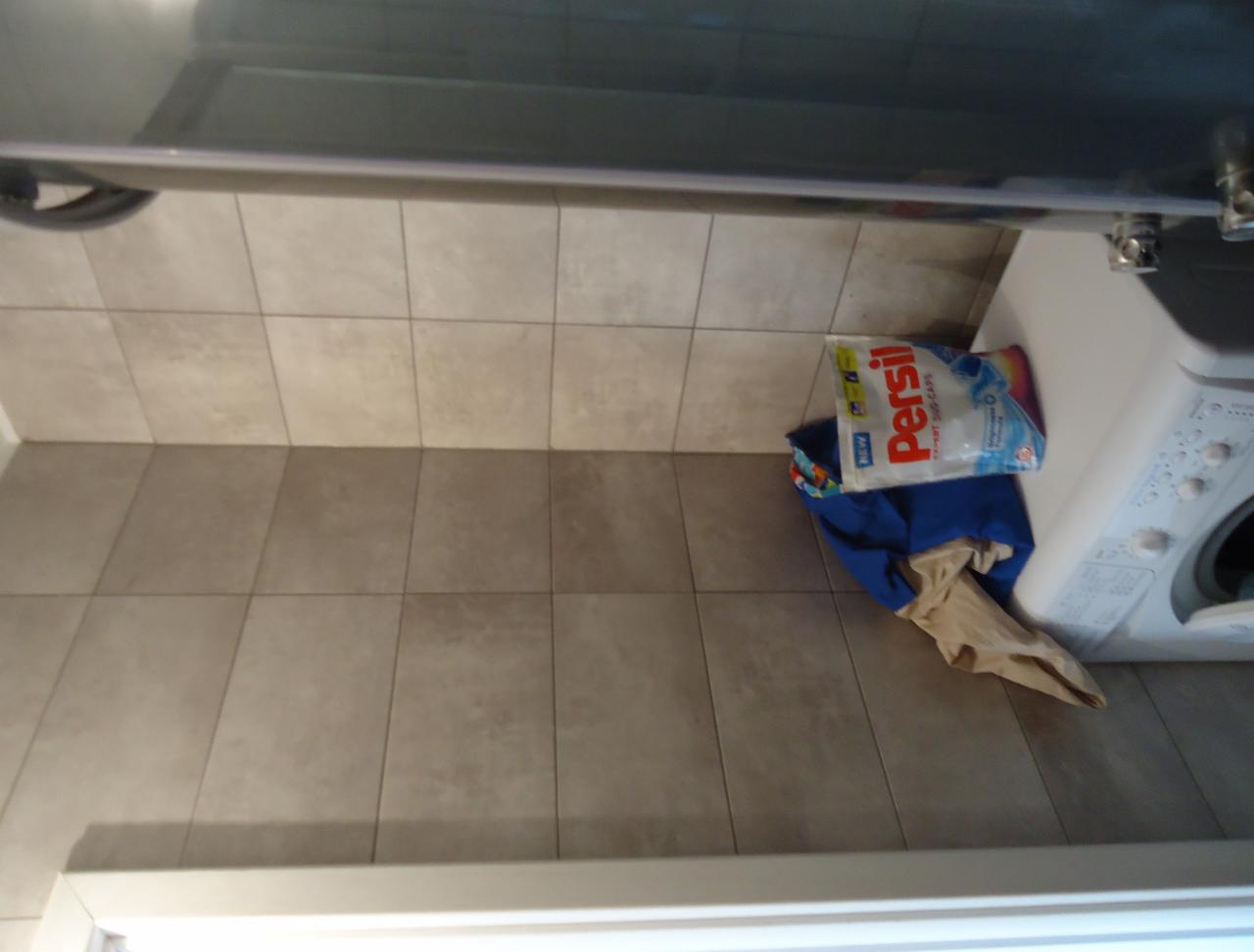 Ułożone płytki podłogowe w łazience, widoczna pralka i proszek do prania. Płytki w odcieniach beżu, matowe. Ujęcie z góry, obrócone o 90 stopni.