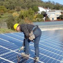Pracownik w kasku montuje panele słoneczne na dachu. W tle drzewa i budynki mieszkalne. Używa wiertarki akumulatorowej. Widok z boku, lekko z góry.