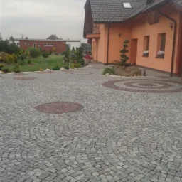 Ułożona szara kostka brukowa na podjeździe domu z elementami dekoracyjnymi z czerwonej kostki, widok na budynek i ogród w tle.