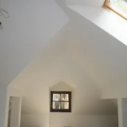 Poddasze po remoncie z oknem dachowym i małym okienkiem w szczycie, widoczne kable zwisające z sufitu, surowe wykończenie ścian na biało.