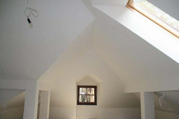 Poddasze po remoncie z oknem dachowym i małym okienkiem w szczycie, widoczne kable zwisające z sufitu, surowe wykończenie ścian na biało.