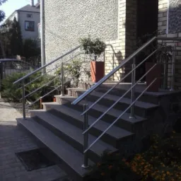 Schody wejściowe z szarego granitu, zabezpieczone nowoczesną balustradą ze stali nierdzewnej. Donice z roślinami po bokach. Elewacja budynku w tle.