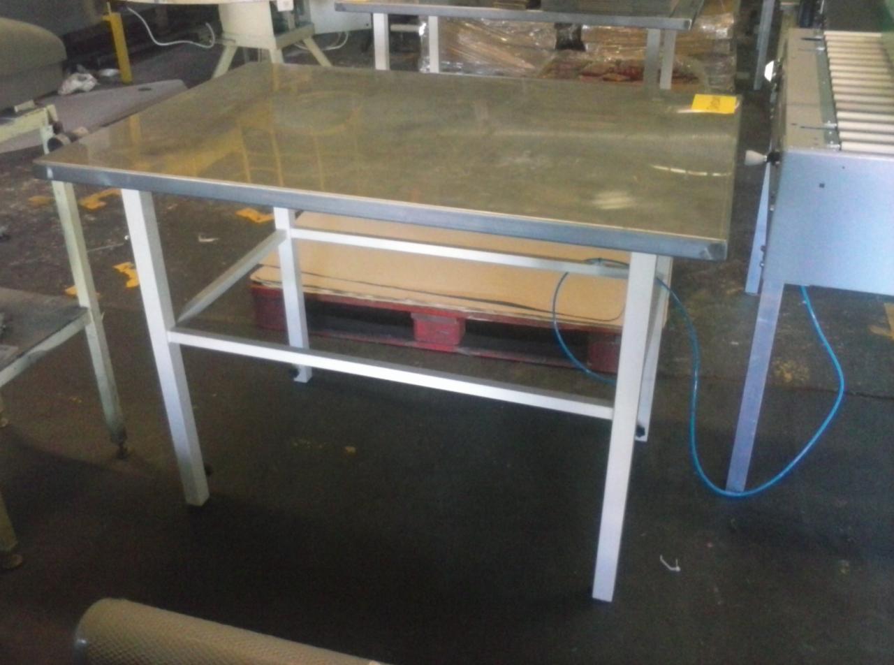 Stół roboczy z metalowym blatem i białymi nogami, z widoczną paletą pod spodem. Ujęcie z perspektywy, ukazujące fragment hali produkcyjnej.