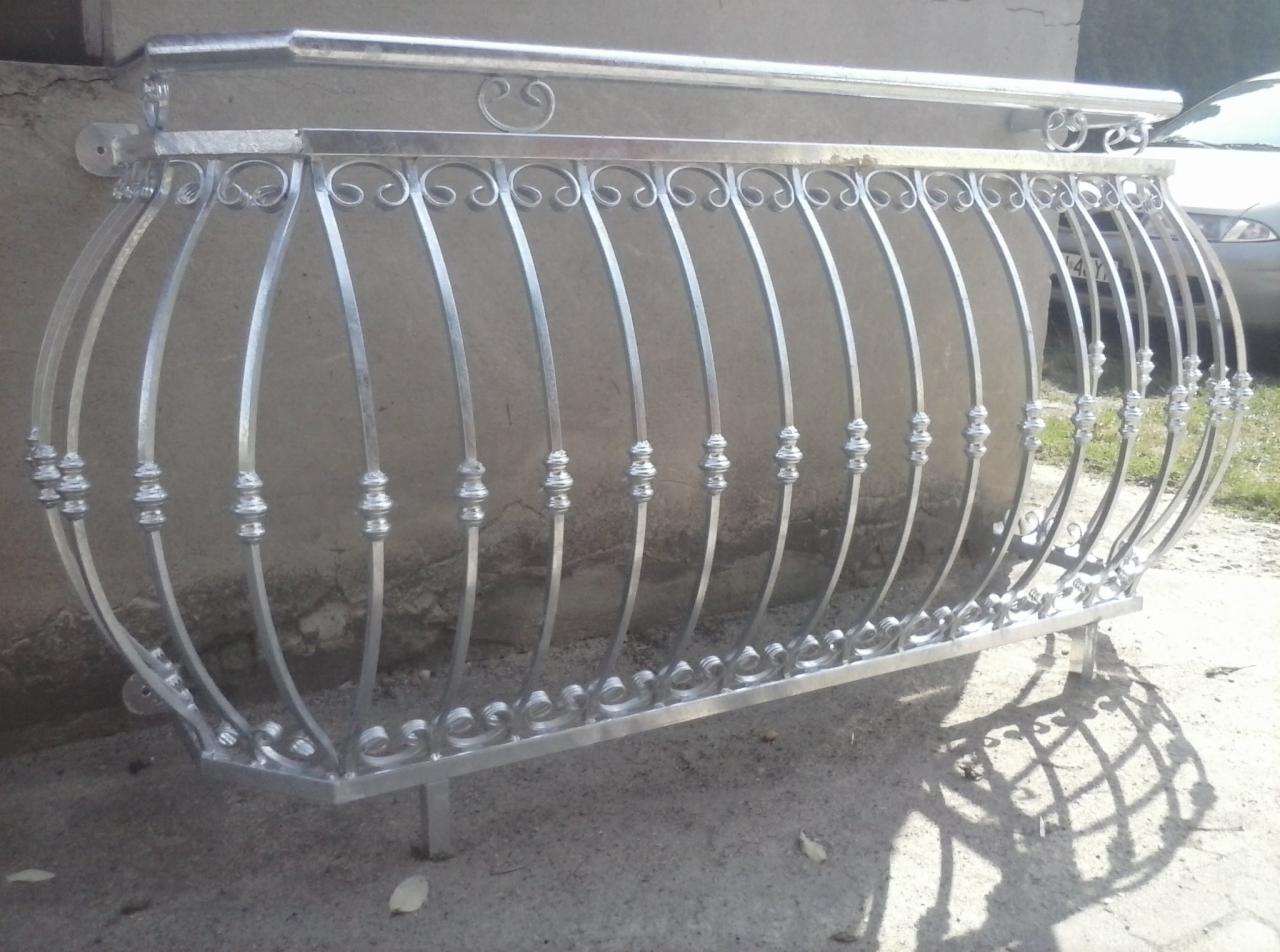 Kuta balustrada balkonowa z ozdobnymi elementami, srebrna, z delikatnymi zdobieniami w kształcie ślimaków i kulek, na tle szarej ściany i fragmentu samochodu.