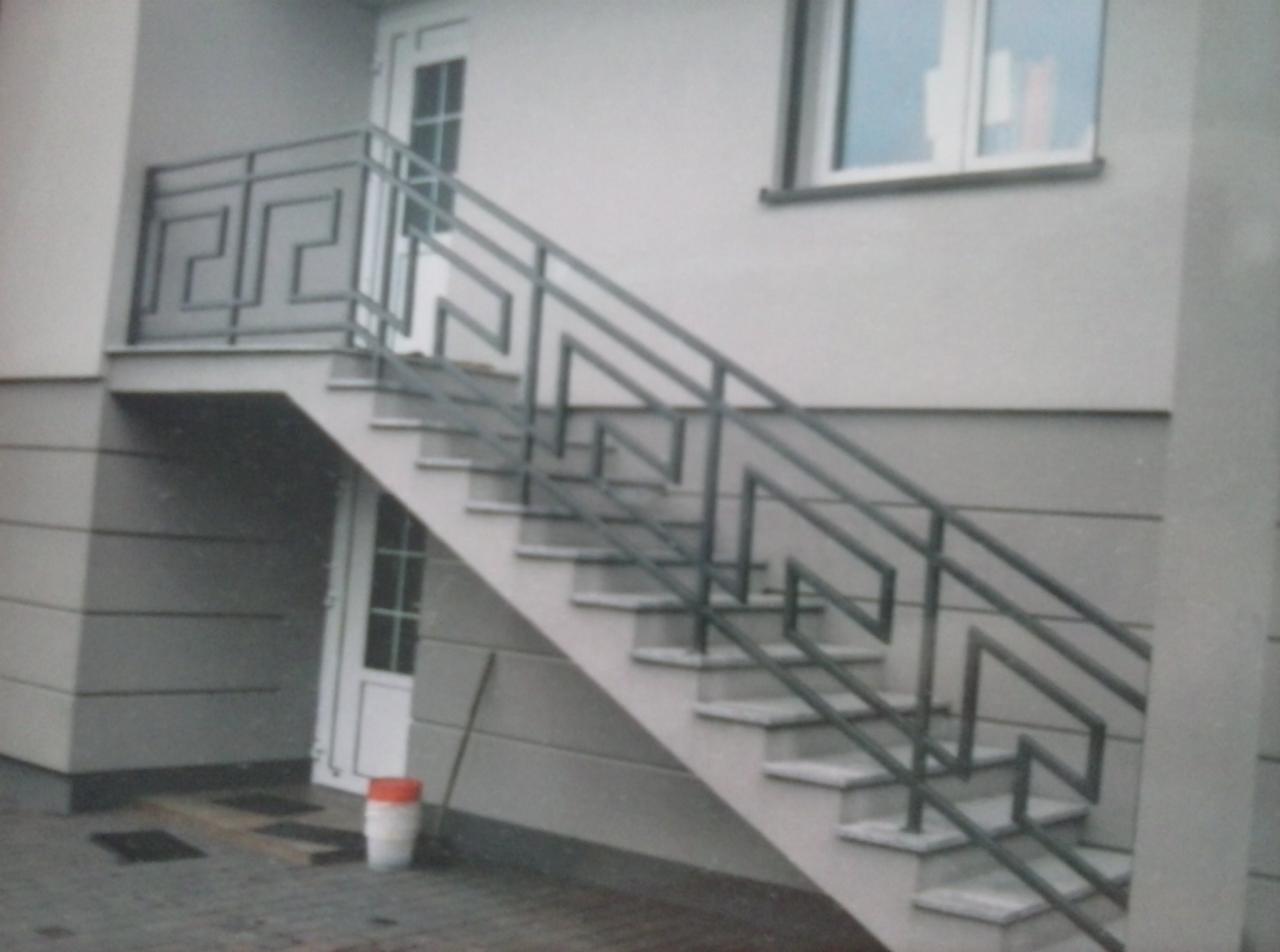 Metalowe schody zewnętrzne z geometrycznymi balustradami w stylu greckim, prowadzące do drzwi. Widoczny fragment elewacji budynku i okno.