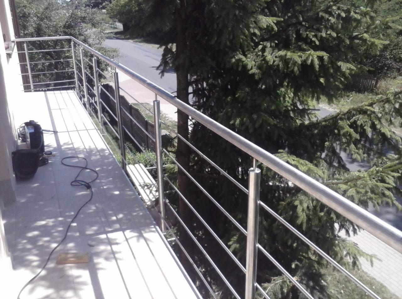 Nowoczesna balustrada balkonowa ze stali nierdzewnej, z widocznym sprzętem montażowym na tle drzew i drogi. Minimalistyczny design, proste linie.