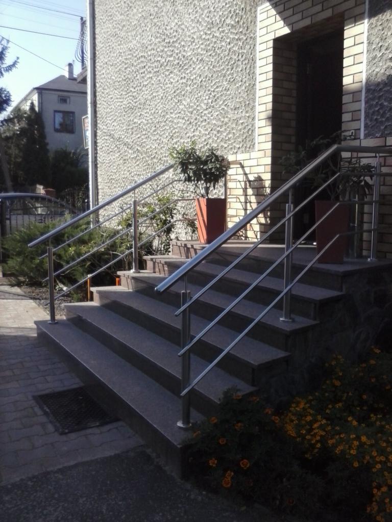 Schody wejściowe z szarego granitu, zabezpieczone nowoczesną balustradą ze stali nierdzewnej. Donice z roślinami po bokach. Elewacja budynku w tle.