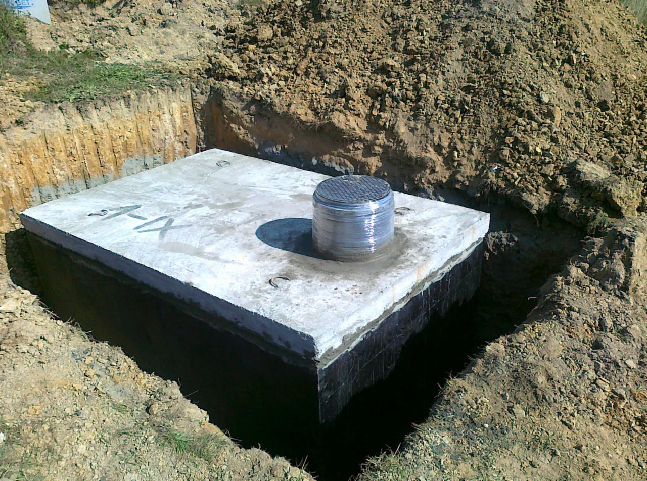 Betonowy zbiornik, prawdopodobnie szambo, umieszczony w wykopie. Widoczna pokrywa zabezpieczona folią. Ściany zbiornika pokryte czarną izolacją.