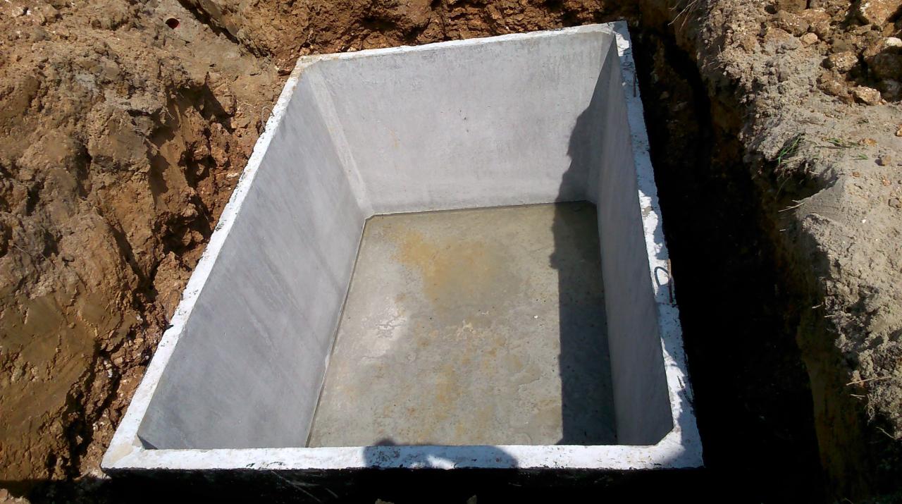 Betonowy zbiornik w ziemi, prawdopodobnie szambo, z widocznymi śladami wody na dnie i uchwytem transportowym. Wykop otoczony ziemią.