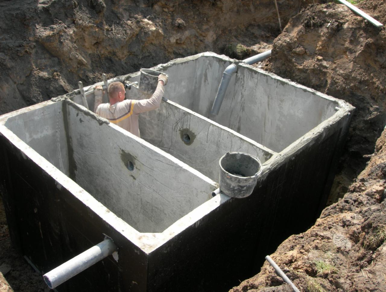 Montaż betonowego zbiornika z przegrodami w wykopie, widoczny pracownik wewnątrz. Rury drenażowe i wiadro na krawędzi. Ujęcie z góry, podkreślające głębokość wykopu.
