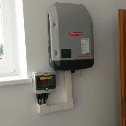 Szary inwerter Fronius SYMO zamontowany na białej ścianie obok okna i skrzynki elektrycznej z widocznymi kablami. W tle fragment drewnianych drzwi.