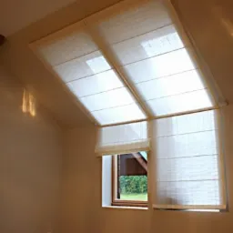 Beżowe rolety rzymskie zamontowane na oknach dachowych i oknie pionowym w jasnym pokoju na poddaszu.